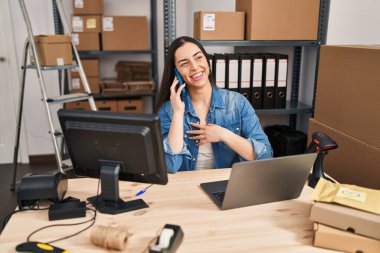 Young beautiful hispanic woman ecommerce business worker using laptop talking on smartphone at office