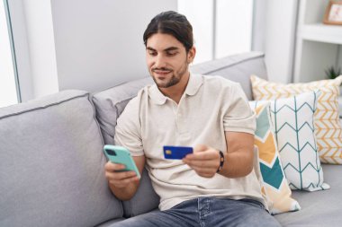 Genç İspanyol adam akıllı telefon ve kredi kartı kullanıyor. Evdeki kanepede oturuyor.