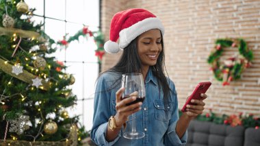 Afrika kökenli Amerikalı kadın akıllı telefon kullanıyor. Evde Noel 'i kutlamak için şarap içiyor.