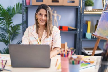 Young hispanic woman artist smiling confident using laptop at art studio