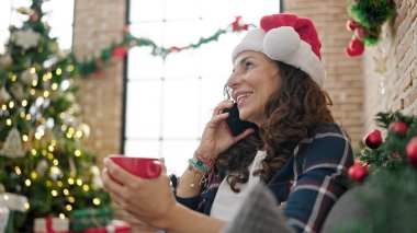 Orta yaşlı İspanyol kadın akıllı telefondan konuşuyor evde Noel 'i kutlamak için kahve içiyor.