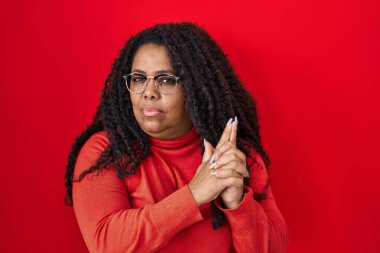 Artı, kırmızı arka planda dikilen boyda İspanyol bir kadın sembolik bir silahla el hareketi yapıyor, cinayet silahlarıyla oynuyor, sinirli bir yüz ifadesi var. 