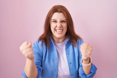 Pembe arka planda kızıl saçlı genç bir İspanyol kadın öfkeyle bağırırken kızgın ve kızgın yumruklarını kaldırıyor. öfke ve agresif konsept. 