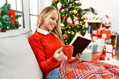 Kafkasyalı genç bir kadın kitap okuyor ve evdeki Noel ağacının yanında kanepede kahve içiyor.