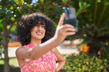 Orta Doğulu genç bir kadın gülümsüyor. Parktaki akıllı telefonun yanında özçekim yapıyor.