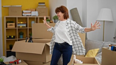 Orta yaşlı bir kadın yeni evinde kollarını açmış kendinden emin bir şekilde gülümsüyor.