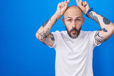 Mavi arka planda dikilen İspanyol dövmeli adam boynuzları gibi parmağıyla komik hareketler yapıyor. 