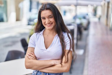 Genç İspanyol kadın gülümsüyor. Kollarını kavuşturup caddede duruyor.