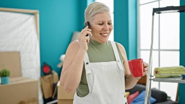 Orta yaşlı, kır saçlı, akıllı telefonla konuşan, yeni evinde kahve içen bir kadın.