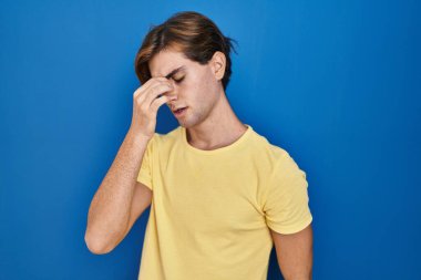 Mavi arka planda duran genç adam yorgun burnu ve gözleri ovuşturuyor yorgunluk ve baş ağrısı hissediyor. Stres ve hüsran kavramı. 