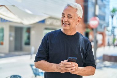 Orta yaşlı, kır saçlı, kendine güvenen, caddede akıllı telefon kullanan bir adam.