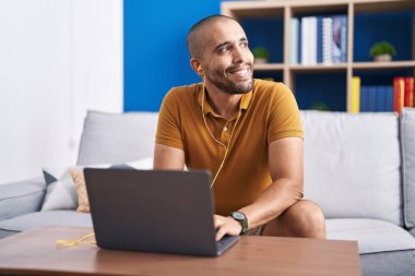 Young latin man using laptop and headphones sitting on sofa at home