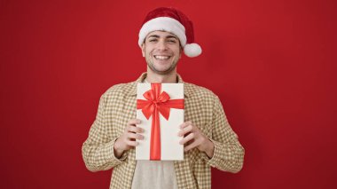 Genç İspanyol adam, kendine güveni tam, Noel şapkası takıyor ve kırmızı arka planda hediyesini tutuyor.