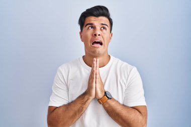 Hispanic man standing over blue background begging and praying with hands together with hope expression on face very emotional and worried. begging. 