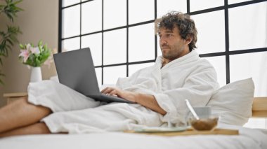 Yatak odasında dizüstü bilgisayar kullanan İspanyol asıllı genç bir adam.