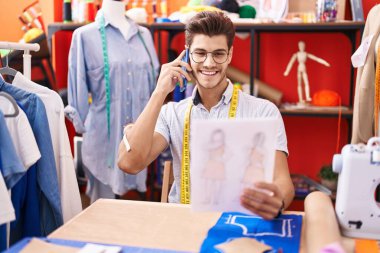 Young hispanic man tailor talking on smartphone looking clothing design at atelier