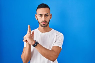 Young hispanic man standing over blue background holding symbolic gun with hand gesture, playing killing shooting weapons, angry face 