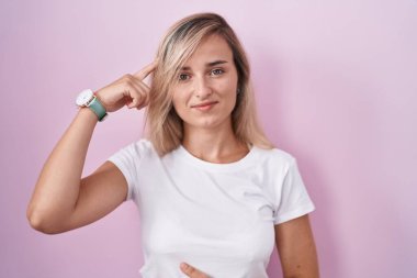 Young blonde woman standing over pink background smiling pointing to head with one finger, great idea or thought, good memory 