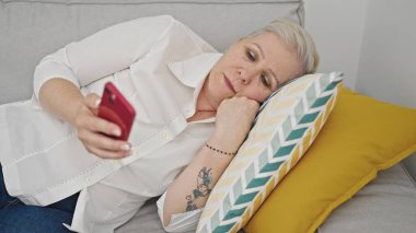 Evdeki kanepede uzanmış akıllı telefon kullanan orta yaşlı gri saçlı kadın.