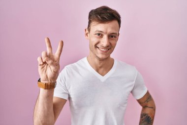 Pembe arka planda duran beyaz adam kameraya bakıp zafer işareti yapıyor. İki numara.. 