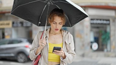 Sokakta şemsiye tutan genç bir kadın akıllı telefon kullanıyor.