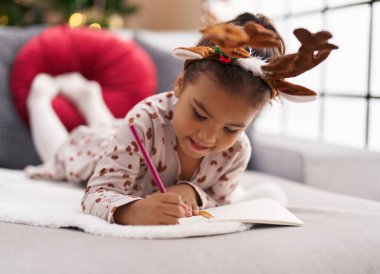Adorable hispanic girl drawing on notebook lying on sofa by christmas tree at home