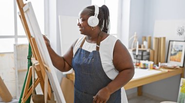 Kendine güvenen Afrikalı Amerikalı kadın sanatçı tutkuyla çiziyor, sanat stüdyosunda müzik dinliyor, kıvrımlarını kucaklıyor ve gülümsüyor.