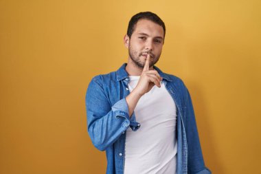 İspanyol adam sarı arka planda dikiliyor ve parmakları dudaklarında sessiz olmak istiyor. sessizlik ve gizli konsept. 