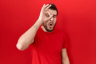 Latin asıllı genç adam, normal kırmızı tişört giyiyor. Şaşırmış yüz ifadesiyle şaşırmış, parmaklarından bakan gözler. İnançsız ifade. 