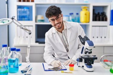 Young hispanic man scientist writing on document holding urine test tube at laboratory