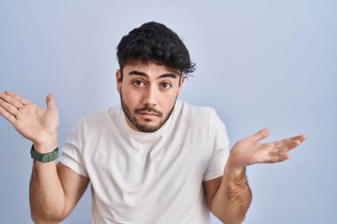 Hispanic man with beard standing over white background clueless and confused expression with arms and hands raised. doubt concept. 