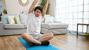 Genç latin adam yerde oturup evde kollarını ve sırtını gererek yoga eğitimi alıyor.