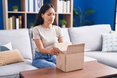 Young hispanic woman opening cardboard box skeptic and nervous, frowning upset because of problem. negative person. 
