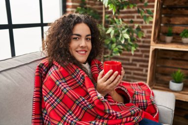 Young beautiful hispanic woman drinking coffee covering with blanket at home