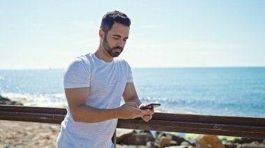 Genç İspanyol adam deniz kenarında akıllı telefon kullanıyor.