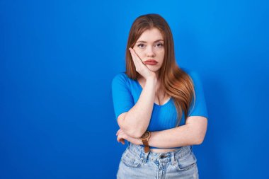 Mavi arka planda duran kızıl saçlı kadın çapraz kollu depresyon sorunlarından yorgun ve sıkılmış göründüğünü düşünüyor.. 
