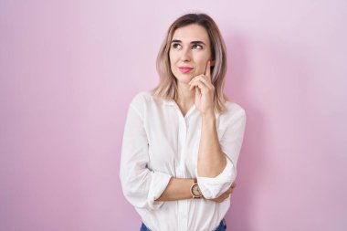 Pembe arka planda duran genç ve güzel bir kadın, elleri çenesinde soru sorarken, düşünceli bir ifade takınıyor. Düşünceli bir yüzle gülümsüyor. şüphe kavramı. 