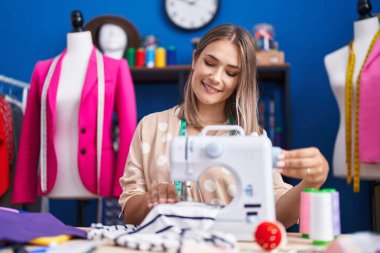 Young caucasian woman tailor smiling confident using sewing machine at sewing studio