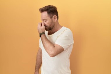 Sarı arka planda duran sakallı orta yaşlı adam yorgun burnu ve gözleri ovuşturup yorgun ve baş ağrısı çekiyor. Stres ve hüsran kavramı. 