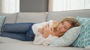 Evdeki kanepede uzanmış akıllı telefon kullanan genç sarışın kadın.