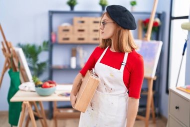 Sanat stüdyosundaki genç kızıl saçlı kadın, elinde bir sanat çantası tutuyor. Tarafsız bir profil çiziyor, doğal bir yüz ifadesi ve kendine güvenen bir gülümseme.. 