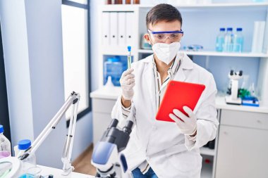 Latin kökenli genç bilim adamı laboratuarda dokunmatik ped kullanarak test tüpü tutuyor.