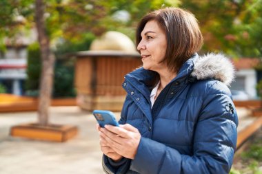 Parkta akıllı telefon kullanan orta yaşlı bir kadın gülümsüyor.