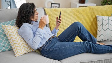 Orta yaşlı İspanyol kadın akıllı telefon kullanıyor evdeki kanepede uzanmış kahve içiyor.