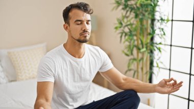 Young hispanic man doing yoga exercise sitting on bed at bedroom