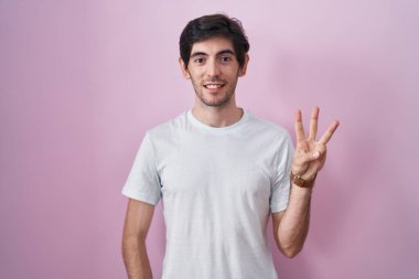 Young hispanic man standing over pink background showing and pointing up with fingers number three while smiling confident and happy. 