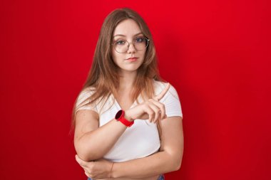 Genç beyaz kadın kırmızı arka planda duruyor. El parmağı yanda, ciddi ve sakin bir yüz ifadesi sergiliyor. 