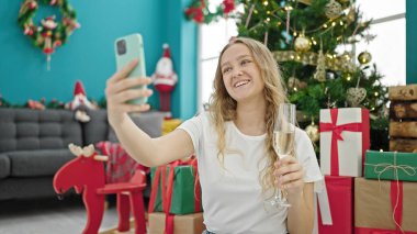Genç sarışın kadın Noel 'i kutluyor. Akıllı telefonuyla selfie çekiyor. Evde şampanya içiyor.