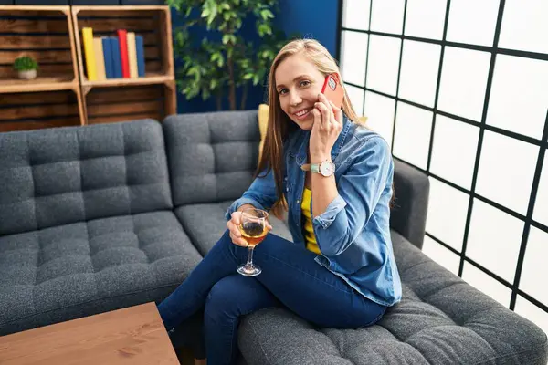 Young blonde woman talking on the smartphone and drinking wine at home