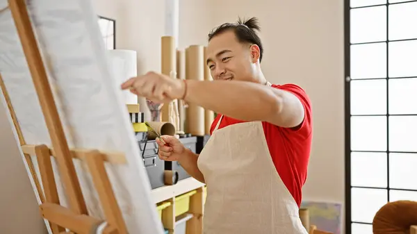 Confident young chinese artist smiling as he enjoys drawing in a vibrant art studio, embodying creativity with a brush and canvas
